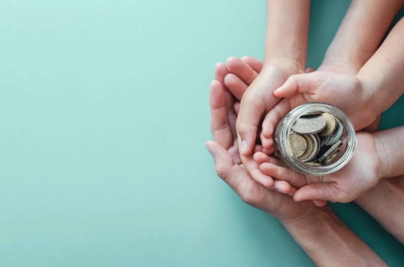 Three pairs of hands, two small and one large, are cupped together holding a glass jar filled with coins against a light blue background, symbolizing saving, sharing, or Accelerated Inheritance Strategies for financial support.