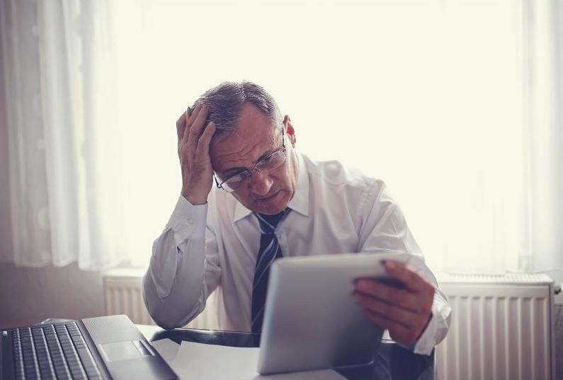 An older man in a shirt and tie sits at a desk, looking stressed as he reviews his tablet. A laptop is open in front of him, suggesting he may be worried about business exit planning mistakes.