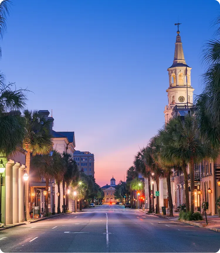 A quiet city street lined with palm trees at dusk, featuring historic buildings and a tall clock tower on the right under a clear, colorful sky.