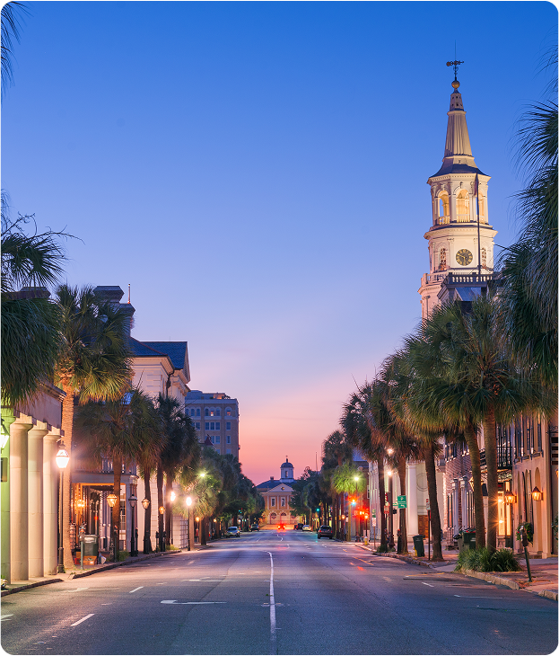 A quiet city street lined with palm trees and historic buildings at dusk, with a tall clock tower on the right and a soft blue and pink sky in the background.