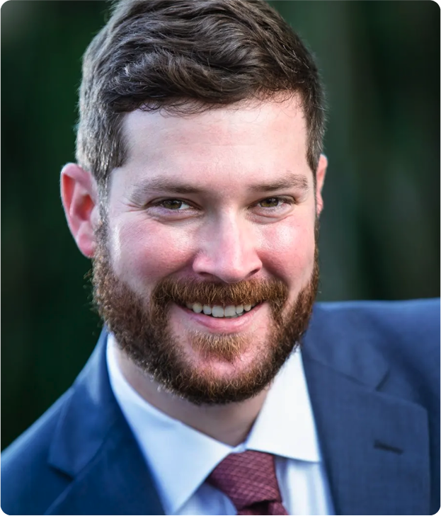 A man with short brown hair and a beard smiles at the camera. He is wearing a blue suit jacket, white dress shirt, and a red tie. The background is blurred and green.