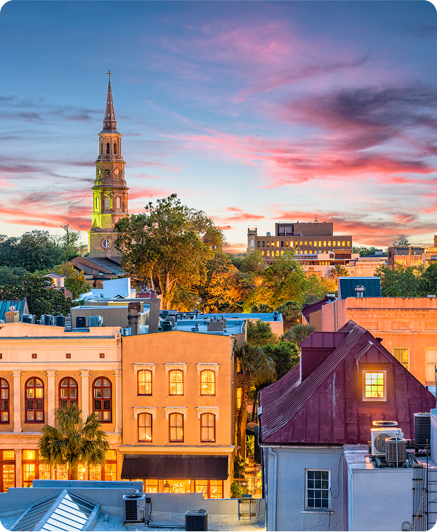 Colorful downtown buildings with lit windows and shops at sunset, with a tall church steeple and trees in the background under a vibrant, partly cloudy sky.