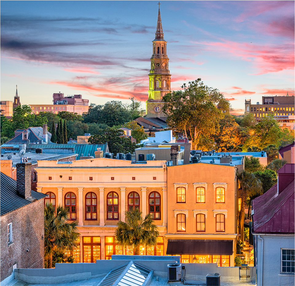 Downtown cityscape at sunset with historic buildings, palm trees, and a tall church steeple rising above the trees; the sky is pink and blue with scattered clouds.