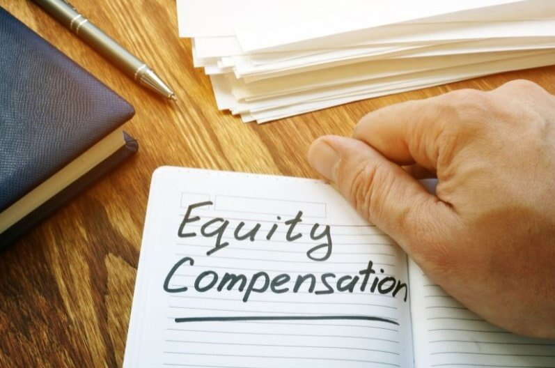 A hand holds open a notebook displaying the handwritten words "Equity Compensation," placed on a wooden desk next to a pen, a closed book, and a stack of papers—perfect for Equity Compensation Financial Planning.