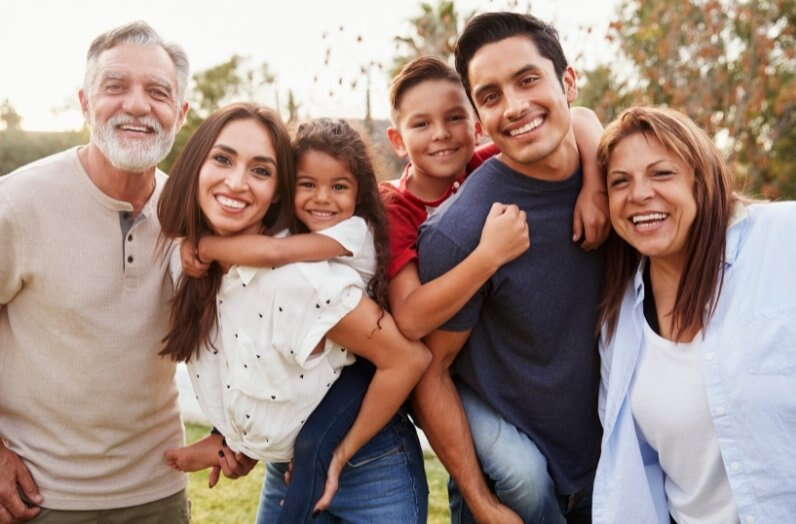 A group of six people, including two children, stand close together outdoors, smiling at the camera. The adults have their arms around each other and the children, capturing a happy family moment—an ideal snapshot for estate planning memories. Trees are visible in the background.