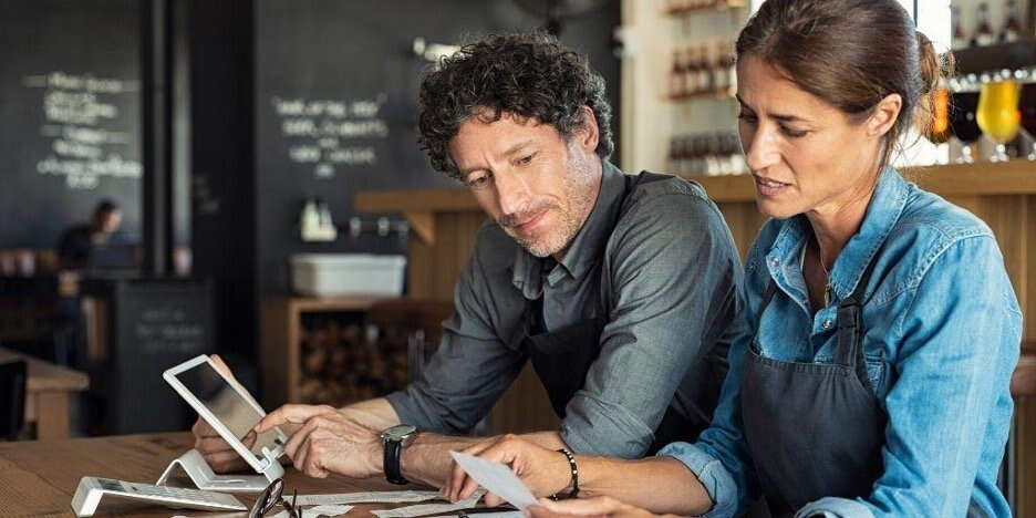 Two people wearing aprons sit at a wooden table, looking at receipts and a tablet. They appear to be working together in a café or restaurant, possibly discussing the best corporate structure for small business financial tasks.