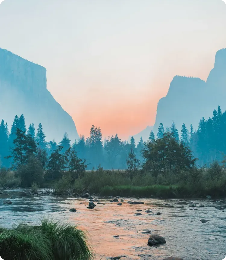 A serene river flows through a lush valley with trees, framed by tall, misty mountains at sunset; soft orange and blue tones fill the sky, creating a peaceful atmosphere.