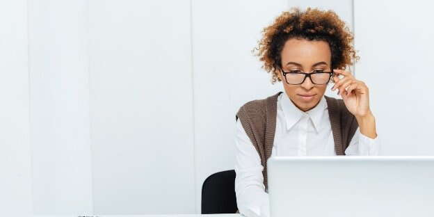 A person with curly hair and glasses sits at a desk, looking focused while working on financial planning for a small business in a bright, minimalist office space.