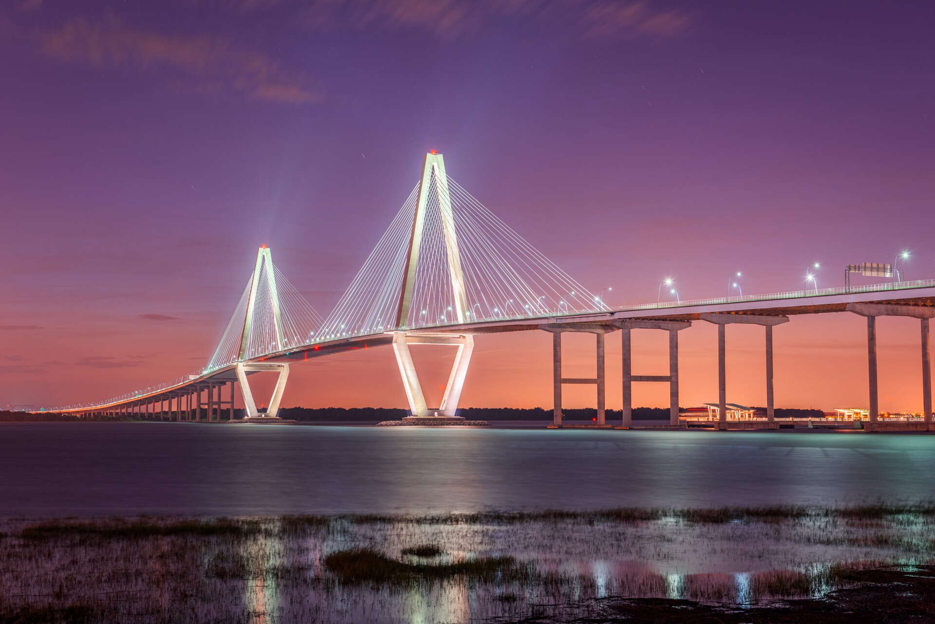 A modern cable-stayed bridge illuminated at dusk stretches across calm water, with glowing lights reflecting on the surface and a purple-orange sky in the background.