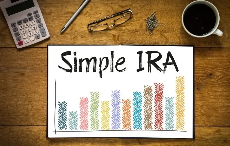 A notebook displaying the words “Simple IRA vs 401(k)” and a colorful bar graph sits on a wooden desk with a calculator, eyeglasses, paperclips, and a cup of coffee nearby.