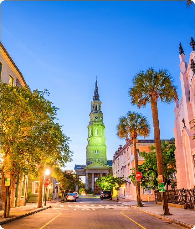 A quiet street in Charleston, South Carolina at dusk, with historic buildings, palm trees, streetlights, and the illuminated steeple of St. Philip’s Church in the background.
