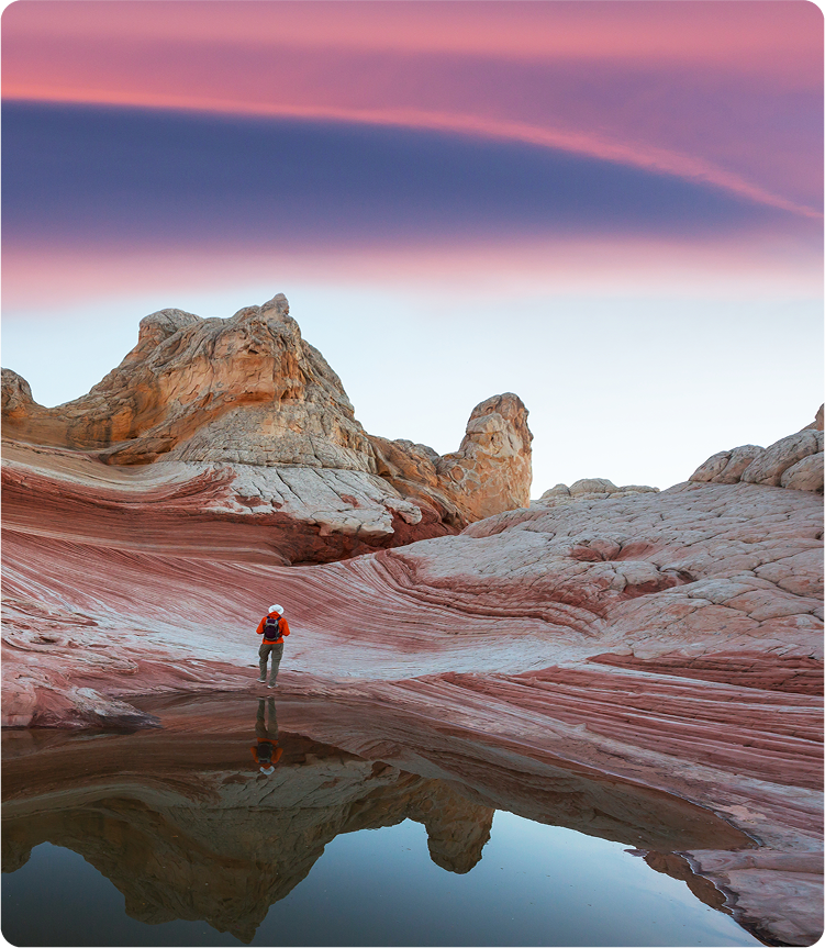 A lone hiker with a red backpack stands on colorful, striped rock formations next to a small reflective pool under a dramatic, pink and purple sunrise sky in a desert landscape.
