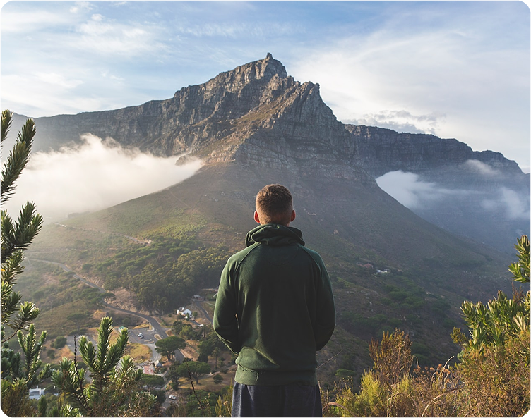 A person in a green hoodie stands facing a scenic mountain landscape, with clouds partially covering the rugged peaks and greenery in the foreground. The sky is mostly clear with some light clouds.