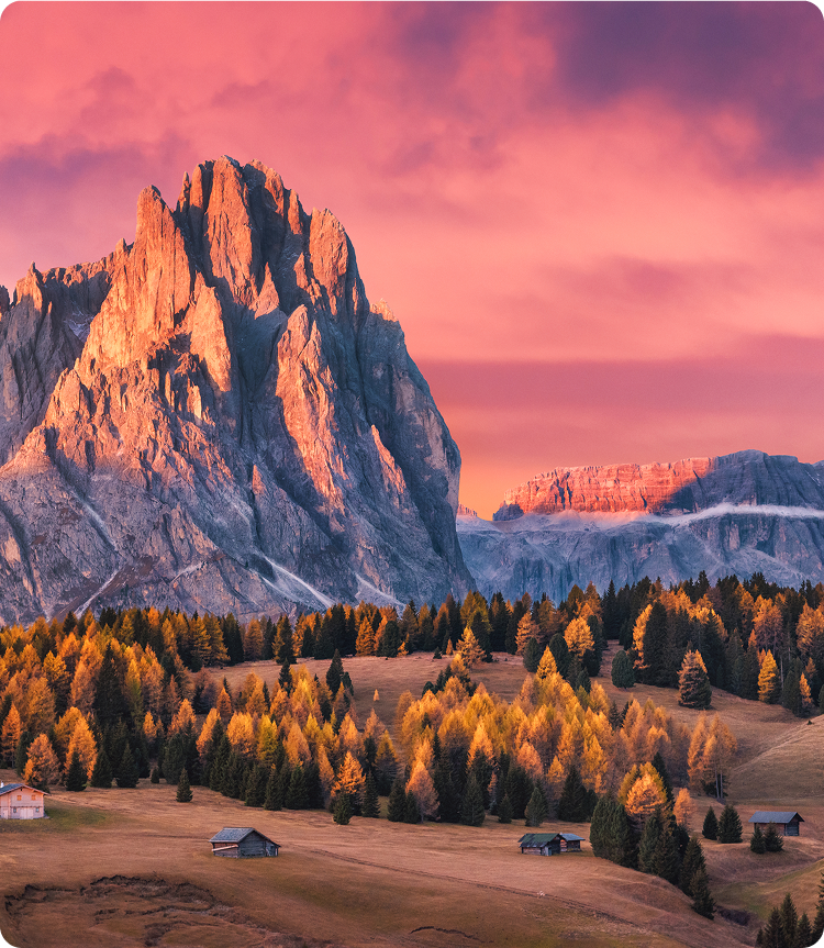 A dramatic mountain peak glows in warm pink and orange light at sunset, overlooking a valley dotted with colorful autumn trees and small wooden cabins under a vibrant sky.