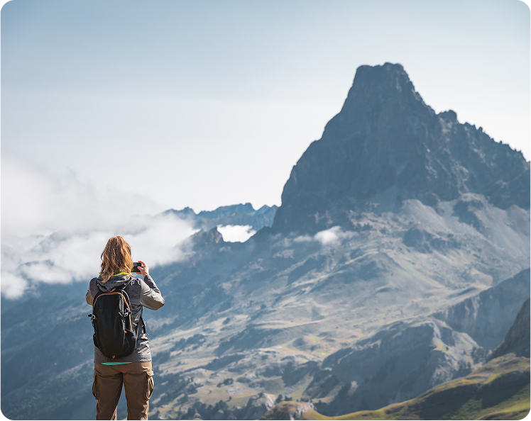 A person with a backpack stands on a grassy hilltop, taking a photo of a tall, rocky mountain peak surrounded by patches of clouds under a clear sky.