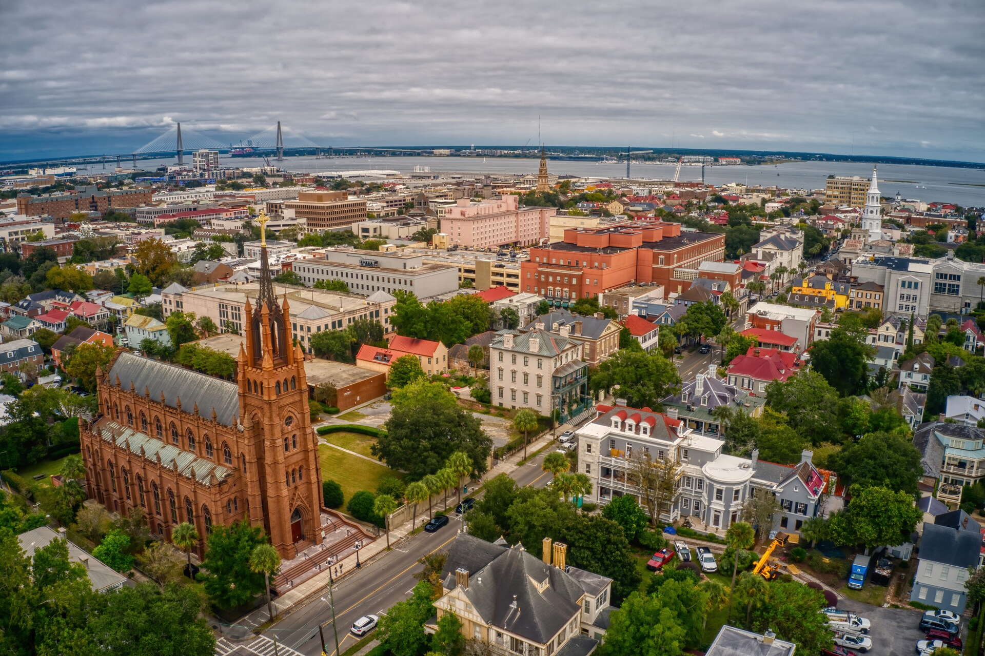 Aerial view of a historic city with a large red-brick church, colorful buildings, tree-lined streets, and a bridge in the distance near a river under cloudy skies.