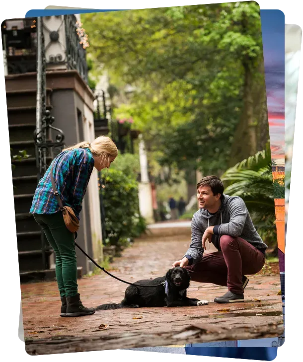A woman stands holding a dog's leash while a man crouches down to pet the black dog on a brick walkway in a leafy, residential neighborhood.