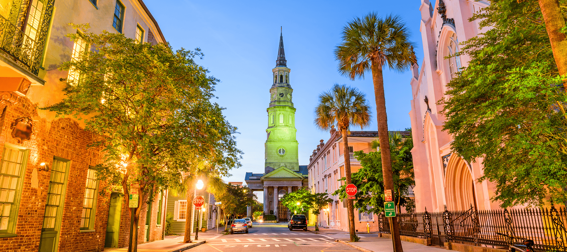 A quiet street in Charleston, South Carolina, lined with historic buildings and palm trees at sunset, features a tall, illuminated church steeple in the background.