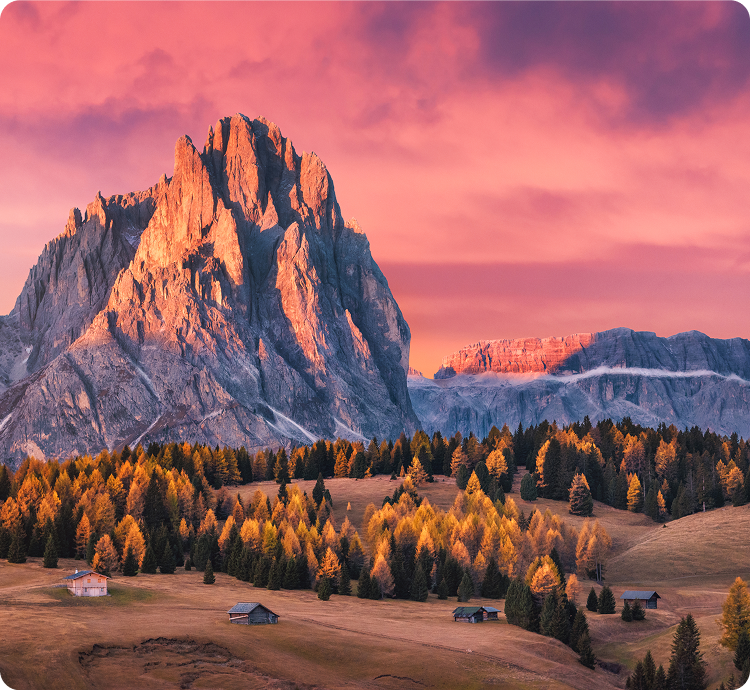A dramatic mountain peak glows under a vibrant pink and orange sunset sky, overlooking a forest of autumn trees and scattered small wooden cabins in a peaceful valley.
