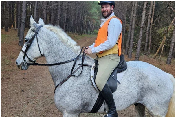 A person wearing a helmet and orange vest is sitting on a gray horse in a forested area with tall trees and a dirt path.