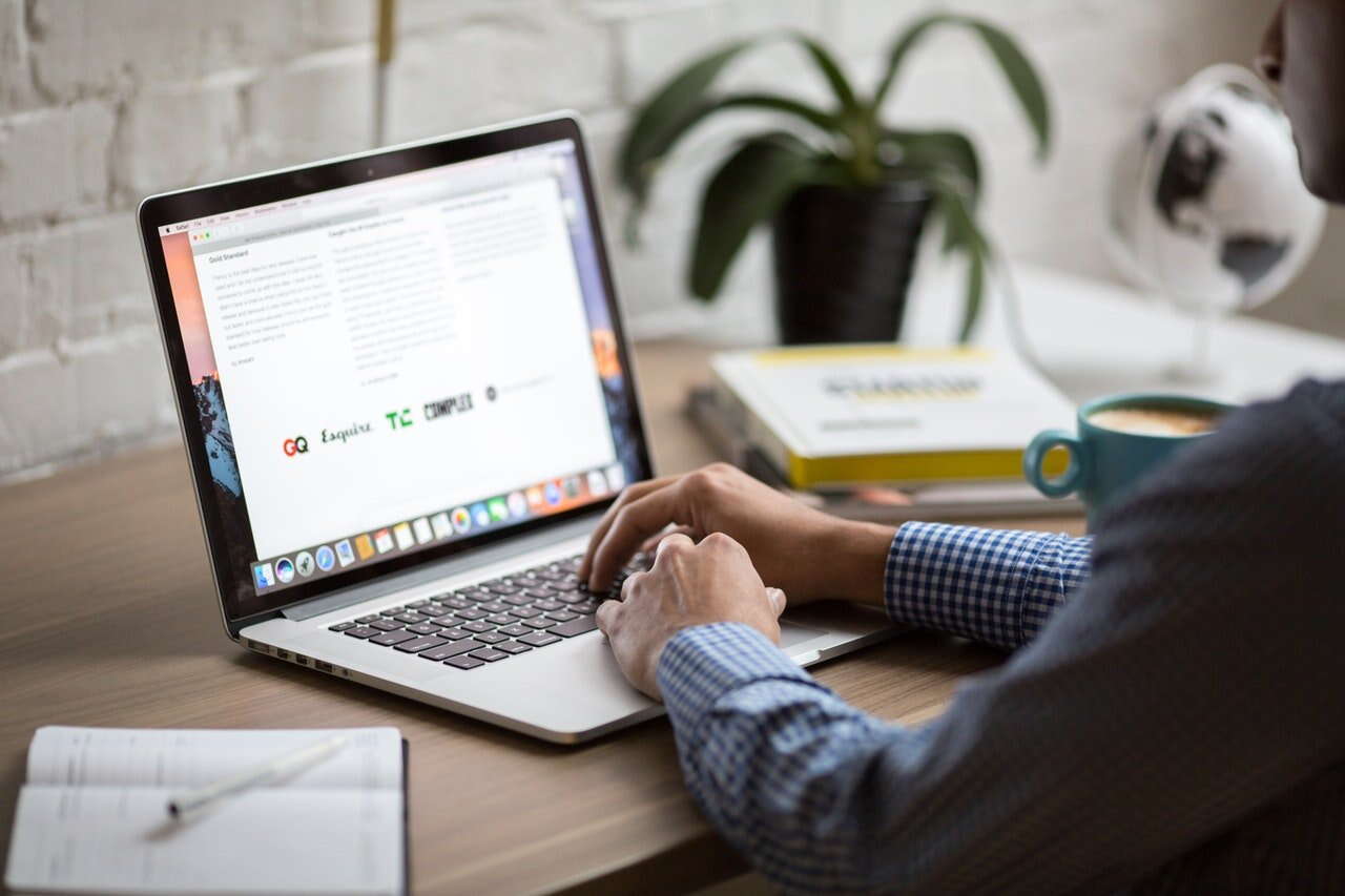 A person in a checkered shirt types on a laptop at a wooden desk with a notebook, pen, potted plant, and a small globe in the background.
