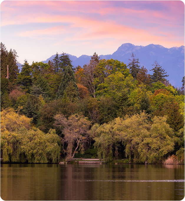 A serene lake surrounded by lush green trees, with distant mountains under a pink and purple sunset sky. The water reflects the trees and sky, creating a peaceful, natural scene.