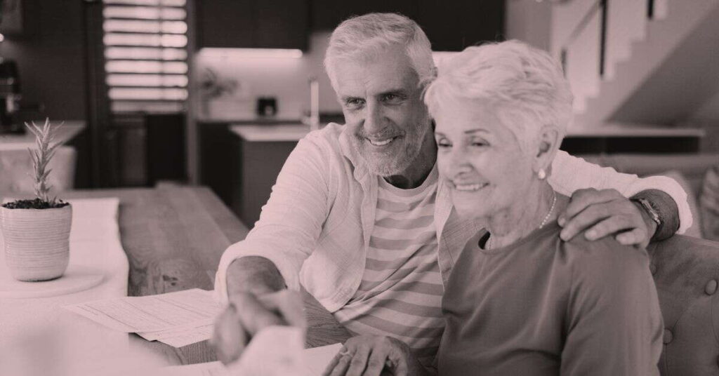An older man and woman sit together at a table, smiling as the man points at papers in front of them—perhaps discussing whether annuities are good for retirement income. The woman leans in closely, and the cozy home setting reflects their comfort and trust.