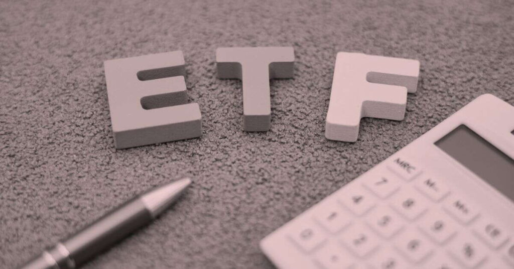 Three large wooden letters spelling "ETF" are placed on a textured surface beside a pen and calculator, suggesting an environment focused on investing in ETFs for long term financial growth.